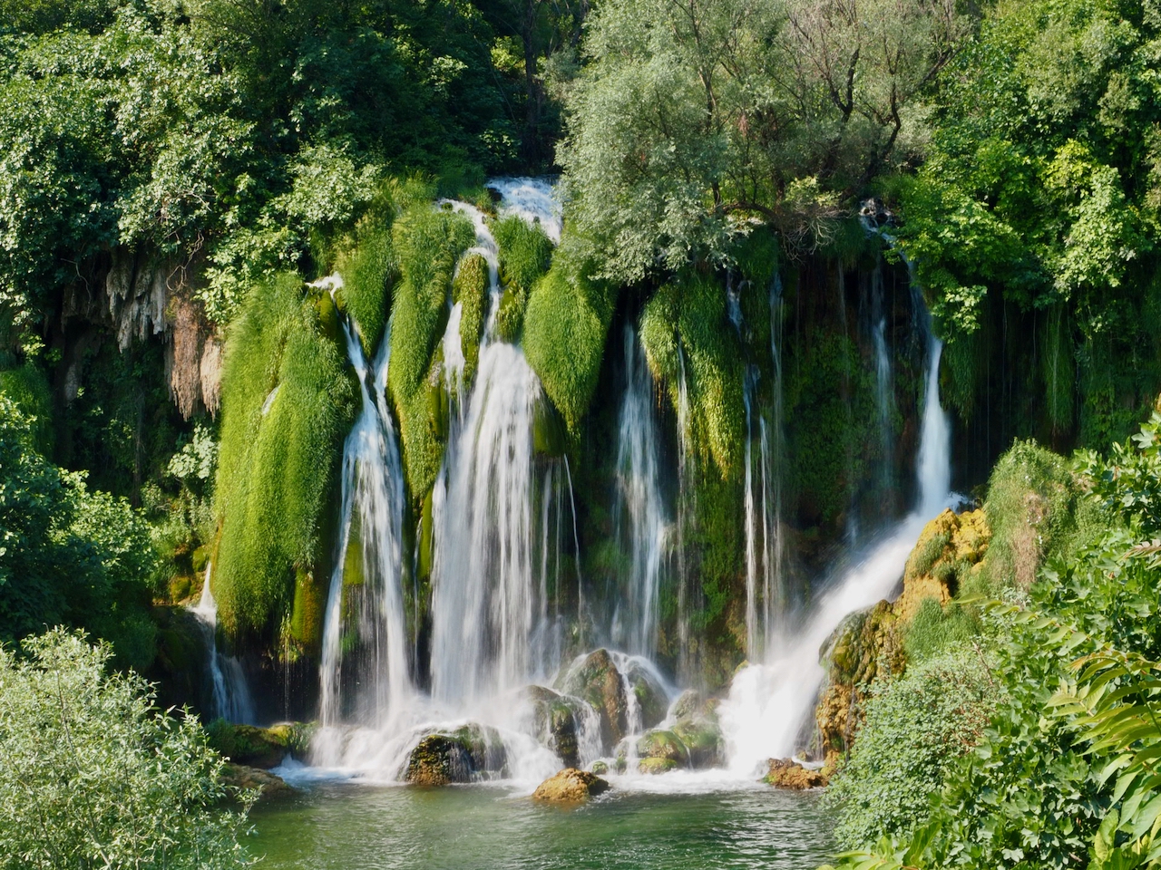 Die breiten Kravica-Wasserfälle fallen über eine grüne Felskante in ein Becken.